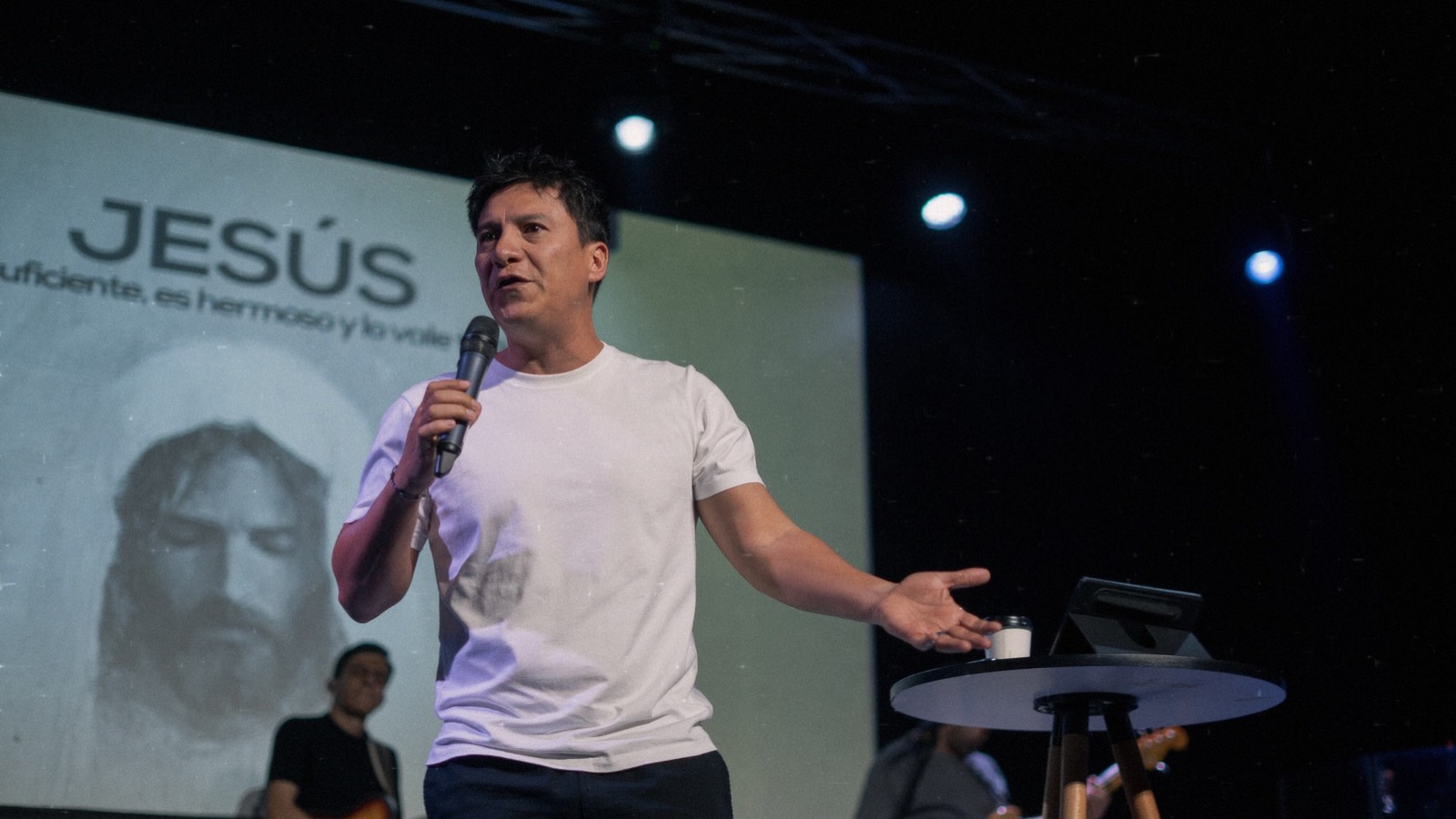 Toño del Río predicando en un escenario; detrás, una pantalla con la palabra JESÚS